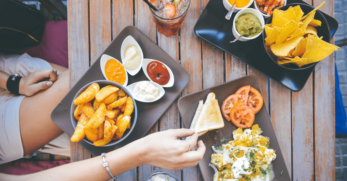 Top view of café-style snack platters with nachos, dips, potato wedges, toast, and fresh vegetables on a wooden table